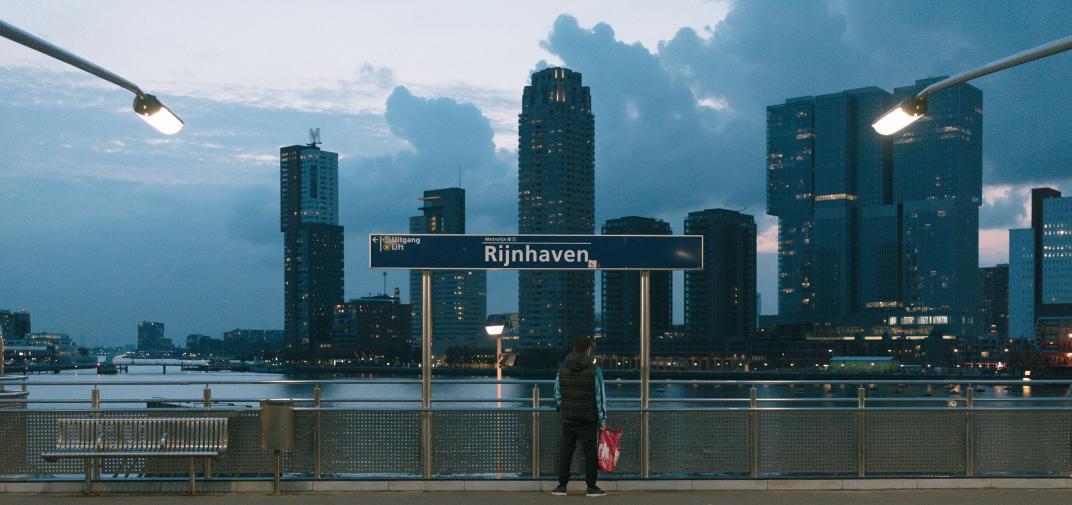 De Kracht van Rotterdam. Feijenoord, 20.00-22.00. Man wacht bij metrostation Rijnhaven. Op de achtergrond hoogbouw op de Kop van Zuid. De foto is gemaakt in opdracht van De Kracht van Rotterdam (DKVR).