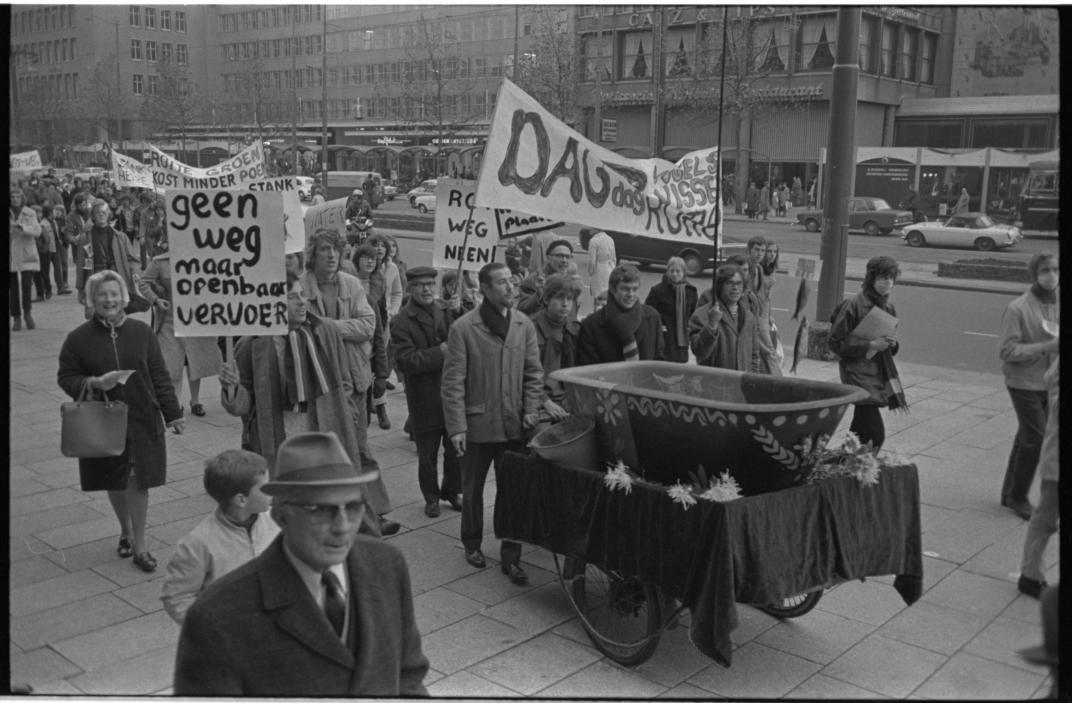  Mensen lopen op de Coolsingel met spandoeken en een baarwagen waarop een beschilderde badkuip is geplaatst.