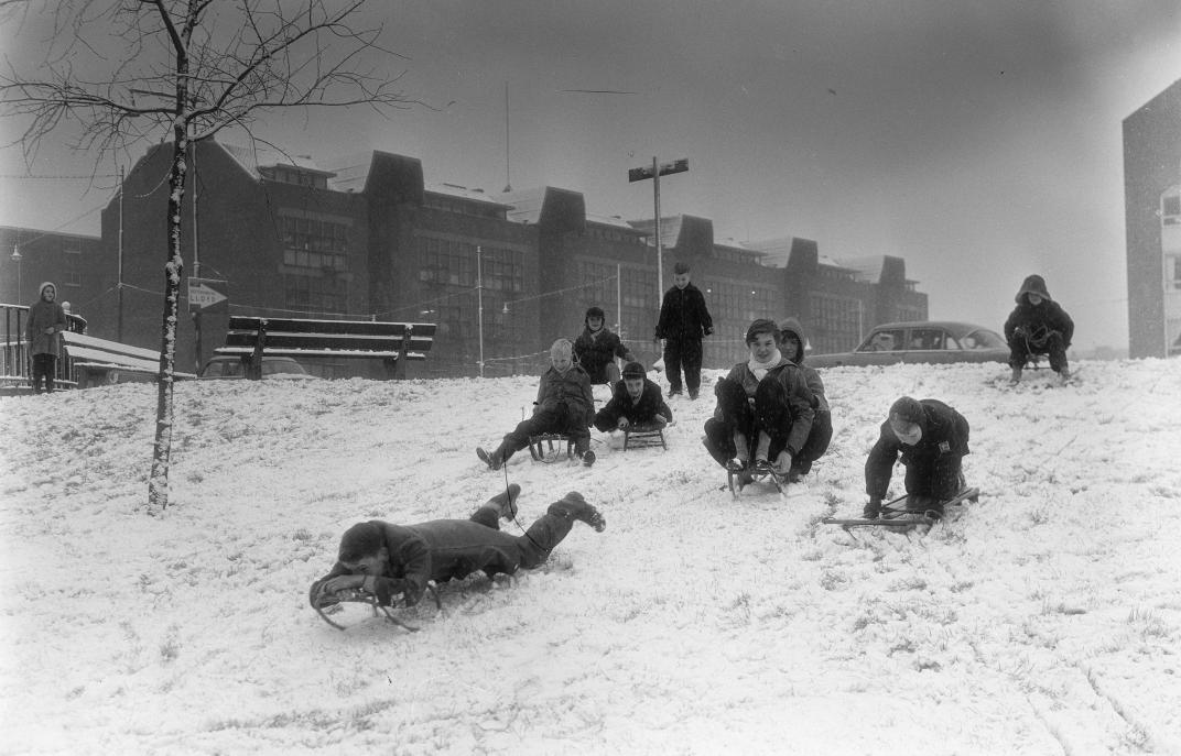Met sleetjes glijden kinderen een besneeuwde dijk af bij de Willem Ruyslaan. Op de achtergrond de Oostzeedijk met Industriegebouw Oostzeedijk.