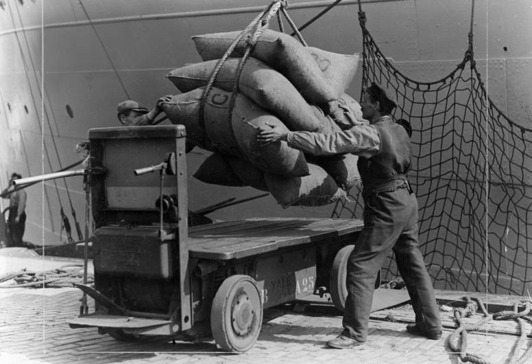 Balen goederen worden vanuit een schip door een havenarbeider op een kar geladen. (1928-1935)