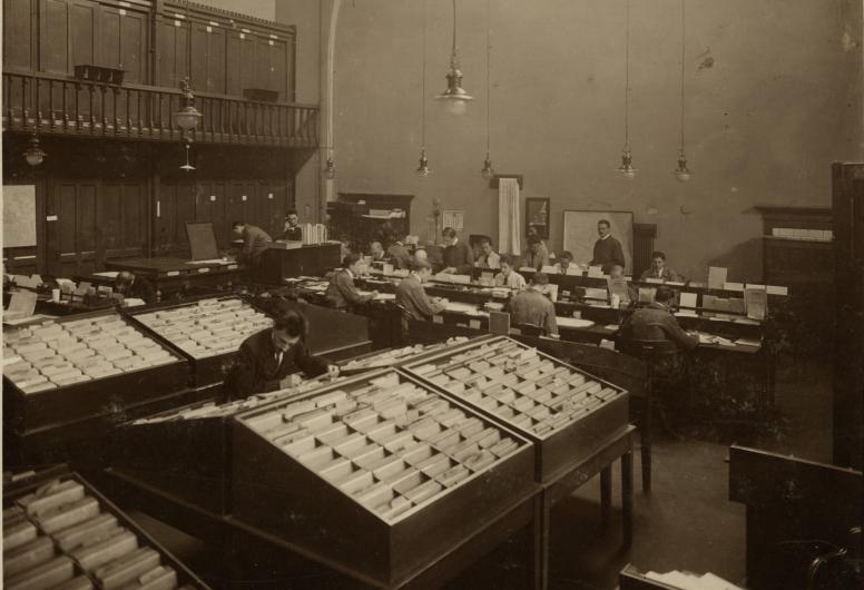 Personeel van de afdeling Bevolking vaan het werk achter bureaus en kaartenbakken met bevolkingsadministratie op het stadhuis aan de Kaasmarkt. 1918 NL-RtSA_4135_XII-30-01-2