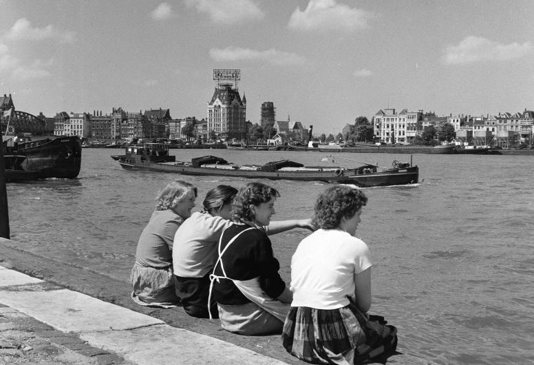 Vier vrouwen zitten op de Maaskade langs de Nieuwe Maas. Een binnenvaartschip vaart voorbij. Op de achtergrond het Witte Huis, rechts de Oosterkade