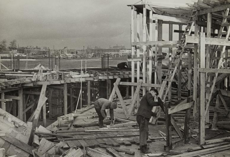 Het Bouwcentrum in aanbouw. Zicht op het houten skelet. Bouwvakkers zijn bezig met stelwerkzaamheden, dec. 1947. Collectie 4100, documentnummer 2016-25-24.