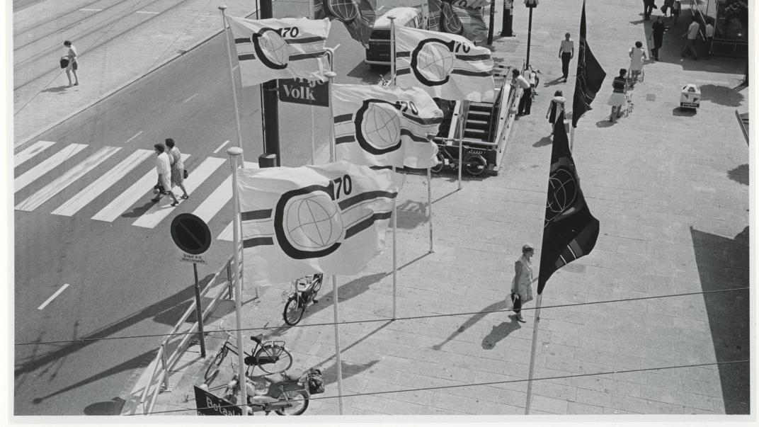 Blik vanuit de kabelbaan op de Coolsingel in zuidelijke richting met vlaggen met het logo van de Manifestatie C70. Fotograaf H.M. Vrijmoet. Collectie 4282, nummer 2142