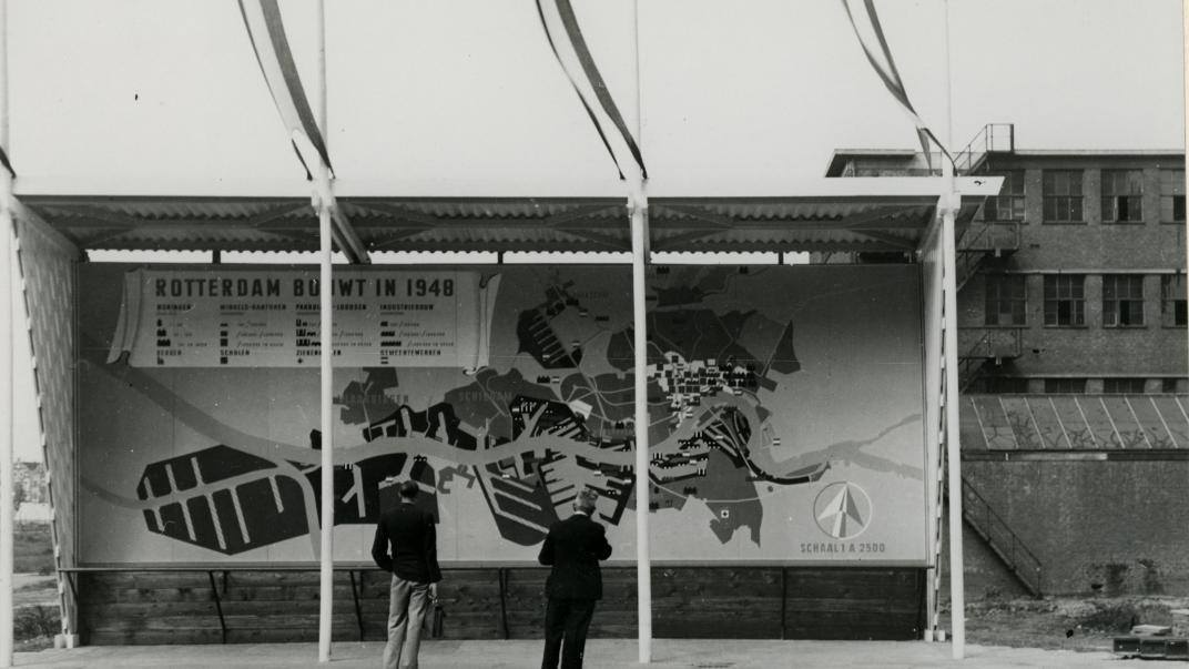 Twee mensen bekijken een groot promotiebord 'Rotterdam Bouwt in 1948' op de Coolsingel. Op de achtergrond rechts een deel van de Ravero garage, 1948. Collectie 4135, XXVI-49-00-00-01