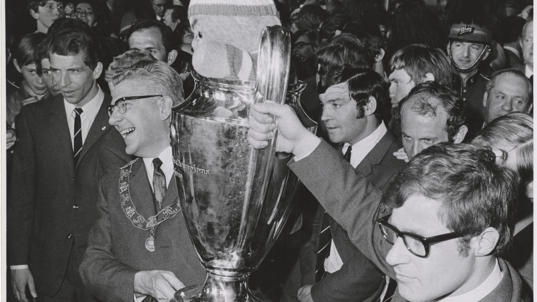 7 mei 1970: Rinus Israël aanvoerder van Feyenoord en burgemeester Thomassen presenteren trots de Europacup I.  Feyenoord is de eerste Nederlandse club die de Europacup I wist te winnen. In de finale versloegen zij, na verlenging, het Schotse Celtic met 2-1. In de cup zit de Celtic-mascotte, een gebreide pop van een halve meter hoog in de clubkleuren groen-wit, die de aanvoerder van de Schotse club cadeau had gedaan aan Feyenoord. Fotograaf Hans Vrijmoet. Collectie 4282, 1970-725-kl